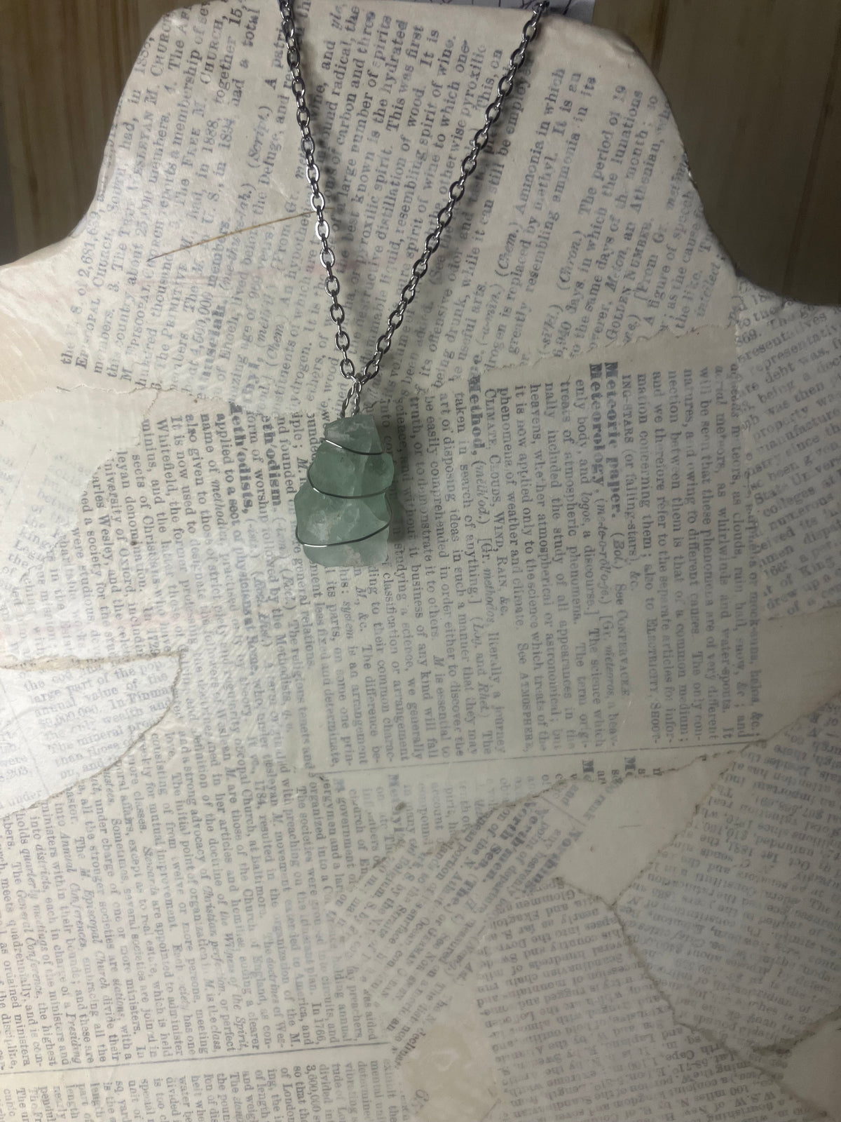 A silver-toned chain necklace with a stacked, organic-shaped light green stone pendant, displayed on a vintage book page mannequin.