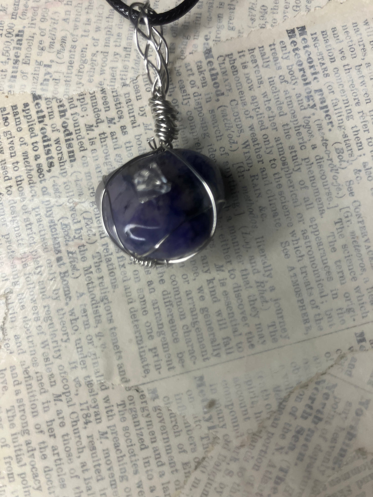 A handmade wire-wrapped pendant featuring a polished dark blue selenite stone, set in silver-toned metal with a twisted wire bail and a black cord, resting on a background of vintage book pages.