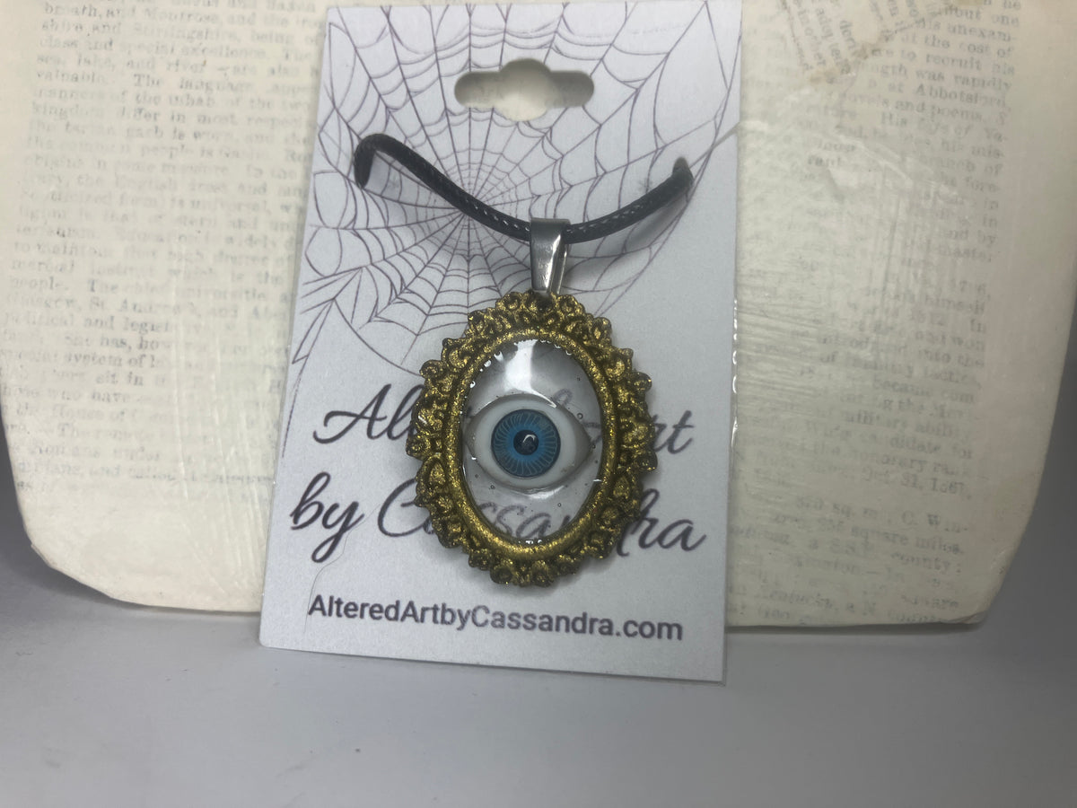A gothic-style evil eye pendant necklace on a black cord, displayed on its white branded card backing. The pendant features a gold filigree frame surrounding a blue and white eye design, set against a background of vintage book pages.