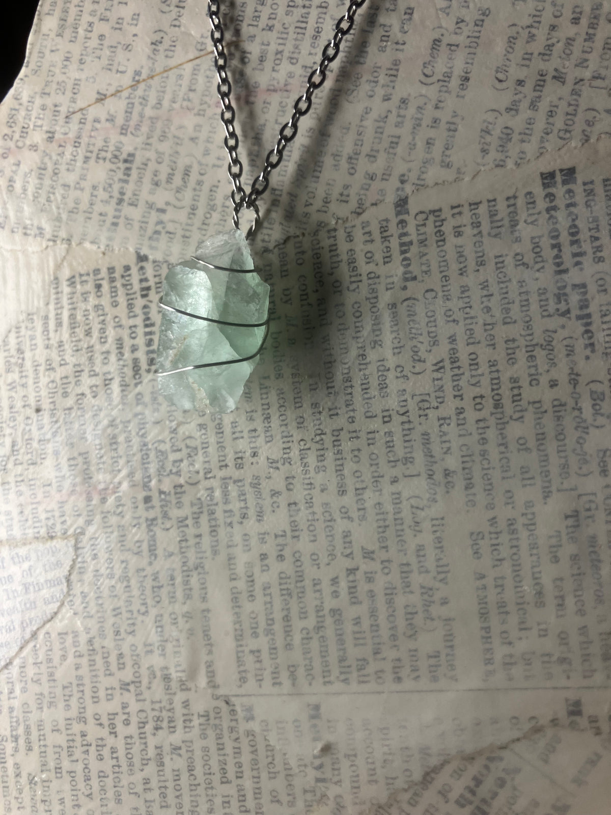 A raw aquamarine pendant wrapped in silver wire, hanging from a silver chain over a vintage newspaper background.